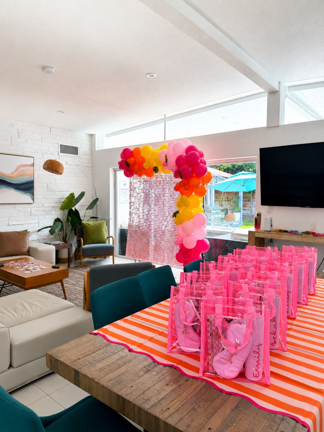 Bright, mid-century living space at the 5BR villa styled for a bachelorette — pink and orange balloon garland, personalized clear tote bags on a striped runner, and a shimmer photo backdrop
